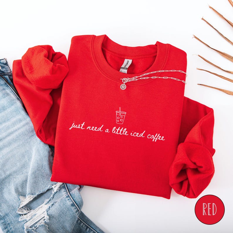 K&ouml;nnte beinhalten: Ein rotes Sweatshirt mit dem wei&szlig;en Schriftzug "just need a little iced coffee". Eine silberne Halskette mit Anh&auml;nger liegt auf dem Sweatshirt. Das Sweatshirt ist mit einer Jeans gefaltet.