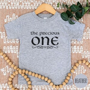 May include: A heather grey short-sleeved t-shirt with the text "the precious one" in black lettering. The text is in a decorative font with a flourish underneath.