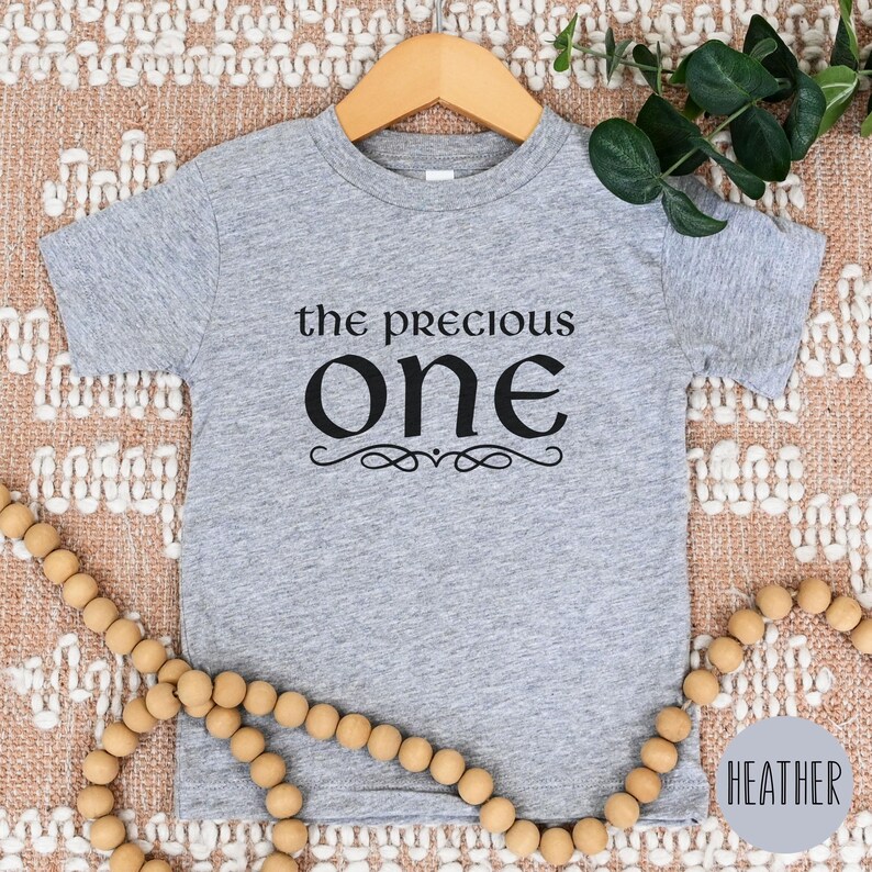 May include: A heather grey short-sleeved t-shirt with the text "the precious one" in black lettering. The text is in a decorative font with a flourish underneath.