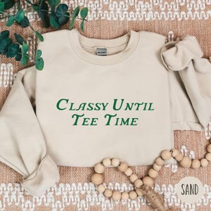 以下が含まれることがあります： 緑色の文字で「Classy Until Tee Time」と書かれたベージュのトレーナー