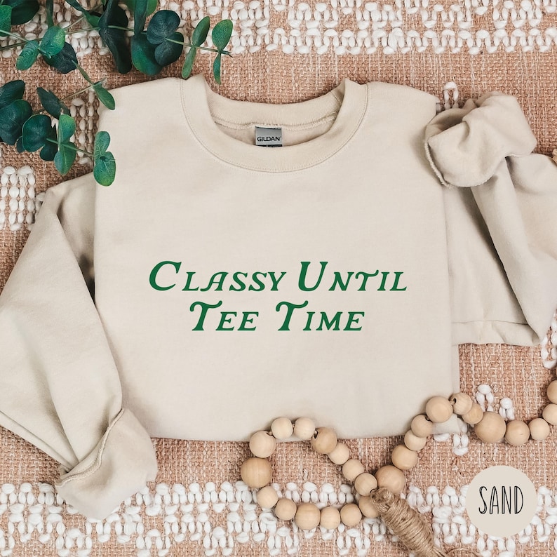 以下が含まれることがあります： 緑色の文字で「Classy Until Tee Time」と書かれたベージュのトレーナー