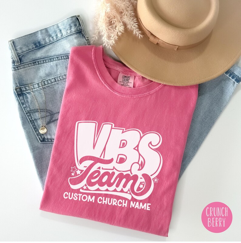 Op de afbeelding: Roze T-shirt met witte tekst die "VBS Team" en "CUSTOM CHURCH NAME" zegt.