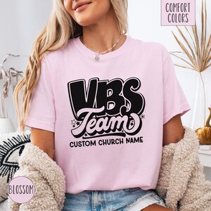 Op de afbeelding: Roze T-shirt met een zwarte afbeelding met de tekst "VBS Team" en "CUSTOM CHURCH NAME".