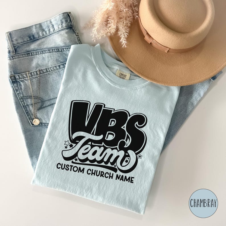 Op de afbeelding: Lichtblauw T-shirt met zwarte tekst die "VBS Team" en "CUSTOM CHURCH NAME" zegt.