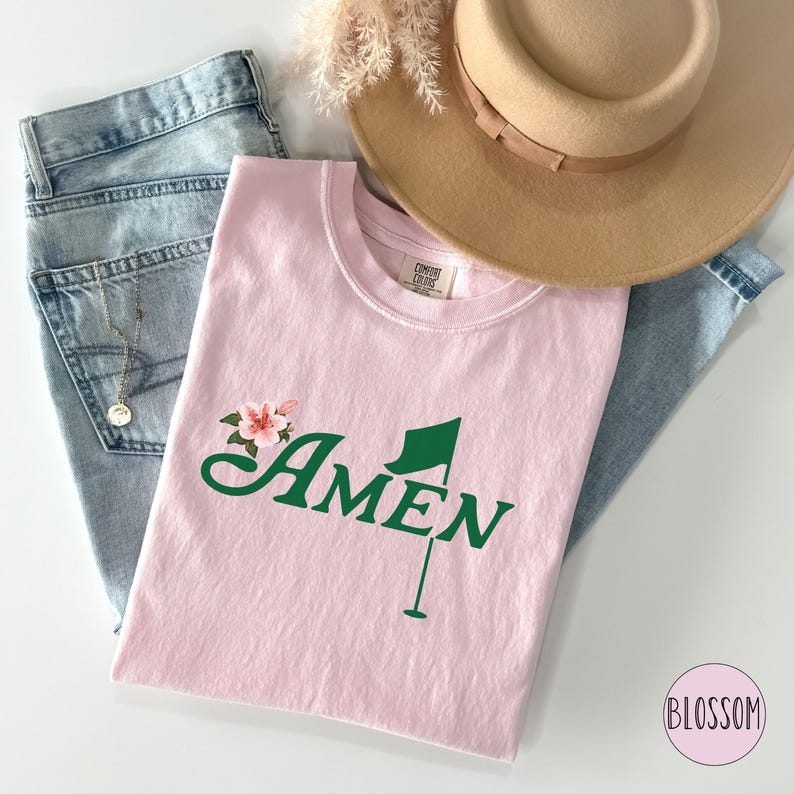 May include: Pink t-shirt with a green golf flag and the word "AMEN" in a floral design.