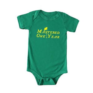 May include: A green baby onesie with short sleeves and a snap closure. The onesie features the text "MASTERED ONE YEAR" in yellow, with a small yellow flag graphic above the word "MASTERED".