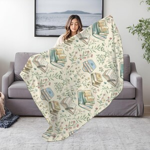 May include: A cream-colored throw blanket with a repeating pattern of watercolor-style illustrations of open books and floral sprigs. The blanket is draped over a gray couch.