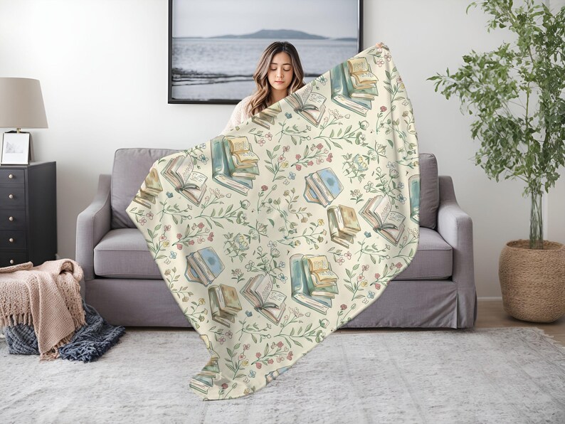 May include: A cream-colored throw blanket with a repeating pattern of watercolor-style illustrations of open books and floral sprigs. The blanket is draped over a gray couch.