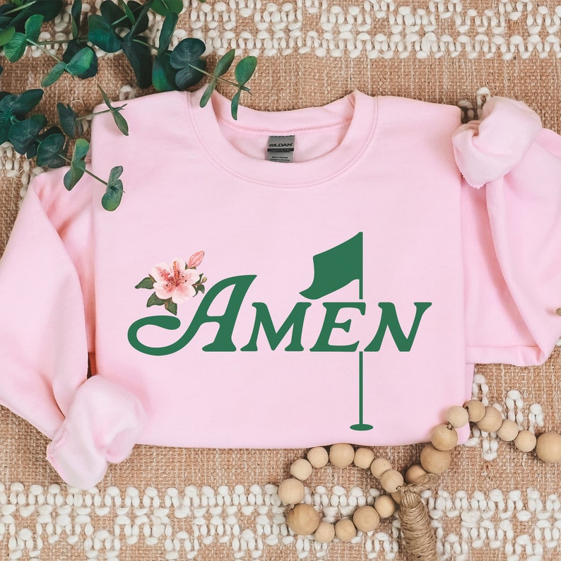 May include: Pink sweatshirt with a green golf flag and the word "AMEN" in a cursive font. A small pink flower is above the word.