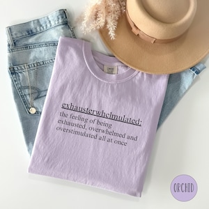 Op de afbeelding: Een lavendelkleurig T-shirt met de tekst "exhausterwhelmulated: the feeling of being exhausted, overwhelmed and overstimulated all at once" in zwart gedrukt.