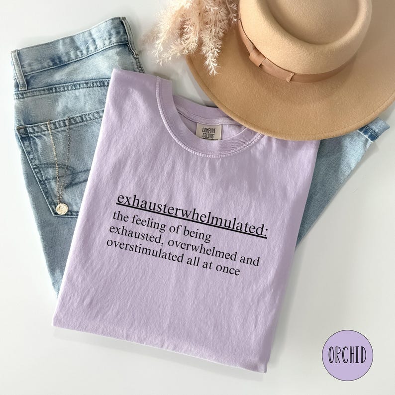 Op de afbeelding: Een lavendelkleurig T-shirt met de tekst "exhausterwhelmulated: the feeling of being exhausted, overwhelmed and overstimulated all at once" in zwart gedrukt.