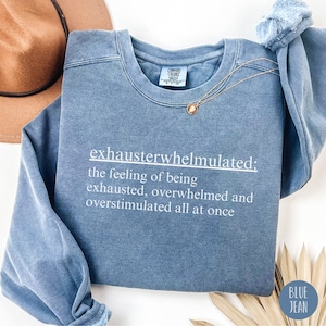 Op de afbeelding: Een blauw sweatshirt met de tekst "exhausterwhelmulated: the feeling of being exhausted, overwhelmed and overstimulated all at once" gedrukt op de voorkant.