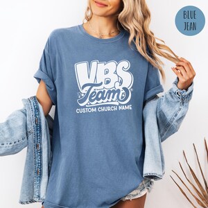 Op de afbeelding: Een jeansblauw T-shirt met witte tekst die "VBS Team" en "CUSTOM CHURCH NAME" zegt.