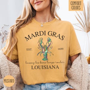 May include: A mustard yellow t-shirt with the text "Mardi Gras" in black lettering. The shirt features a green, gold, and purple lobster illustration with the text "Est. 1699" and "Laissez les bons temps rouler Louisiana".