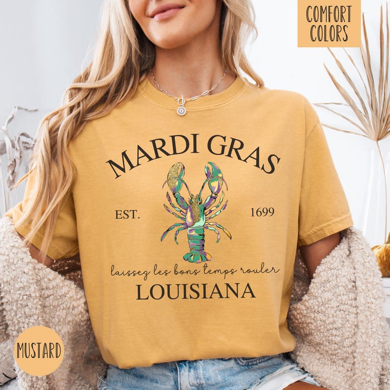 May include: A mustard yellow t-shirt with the text "Mardi Gras" in black lettering. The shirt features a green, gold, and purple lobster illustration with the text "Est. 1699" and "Laissez les bons temps rouler Louisiana".