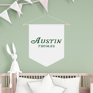 May include: White banner with the name "Austin Thomas" written in green cursive lettering, hanging on a light green wall. A white bunny rabbit figurine is sitting on a white crib with a white pillow.