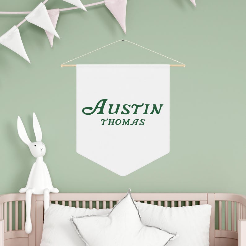 May include: White banner with the name "Austin Thomas" written in green cursive lettering, hanging on a light green wall. A white bunny rabbit figurine is sitting on a white crib with a white pillow.