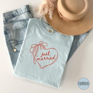 Op de afbeelding: Een lichtblauw t-shirt met een rood hart- en strikdesign, met de woorden "just married". Het shirt wordt gecombineerd met een lichtblauwe jeans en een beige hoed. Een gouden ketting is zichtbaar.
