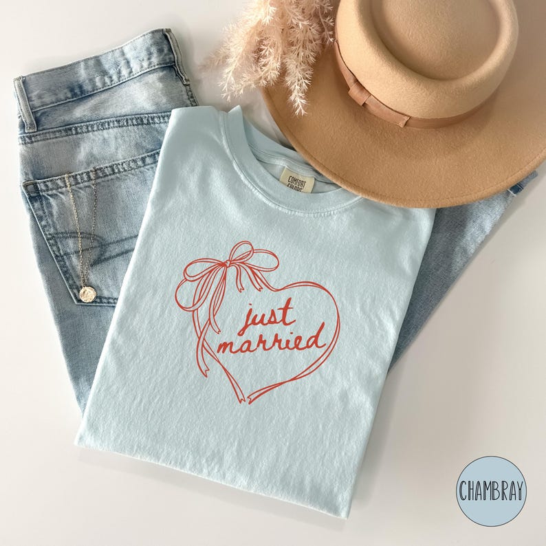 Op de afbeelding: Een lichtblauw t-shirt met een rood hart- en strikdesign, met de woorden "just married". Het shirt wordt gecombineerd met een lichtblauwe jeans en een beige hoed. Een gouden ketting is zichtbaar.