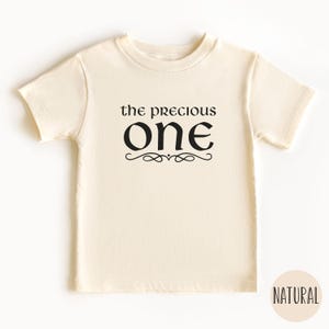 May include: A cream colored short-sleeved t-shirt with the text "the precious one" in black lettering with a decorative flourish.