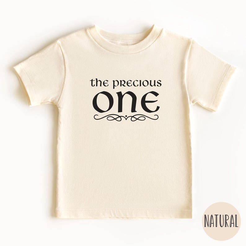 May include: A cream colored short-sleeved t-shirt with the text "the precious one" in black lettering with a decorative flourish.
