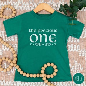 May include: A green short-sleeved t-shirt with the text "the precious one" in white lettering. The shirt is on a light brown background with wooden beads.