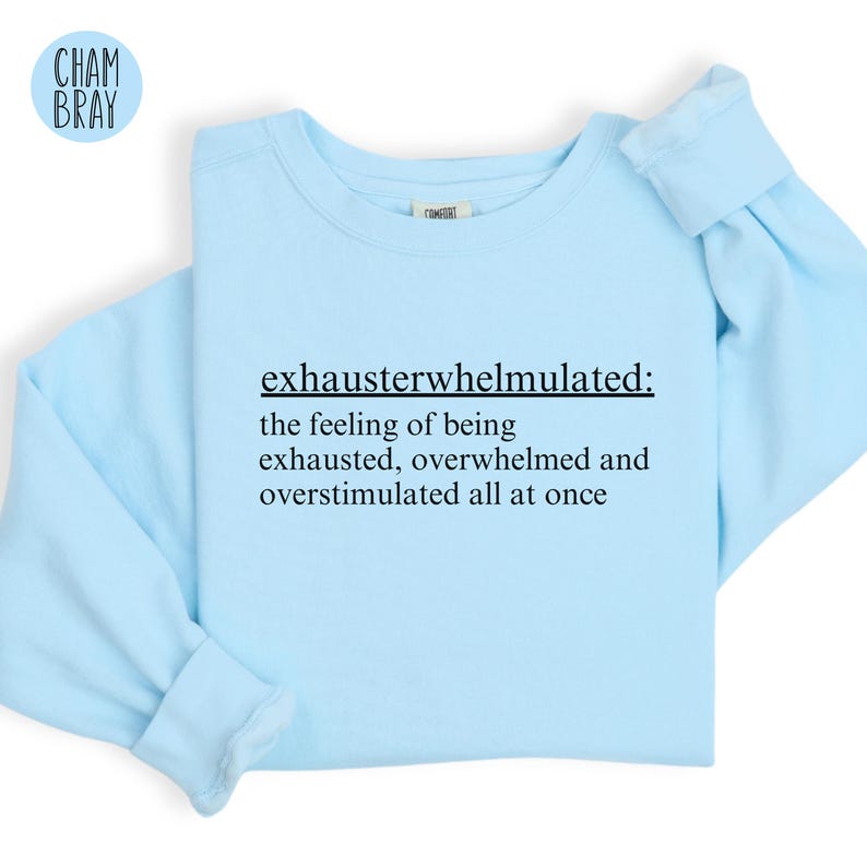 Op de afbeelding: Lichtblauwe trui met de tekst "exhausterwhelmulated: the feeling of being exhausted, overwhelmed and overstimulated all at once" in het zwart gedrukt.