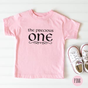 May include: Pink t-shirt with black text that reads "the precious one" with a decorative flourish. The shirt is on a white wood background.