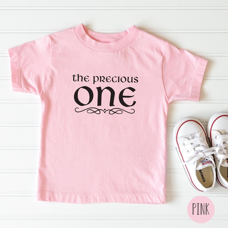 May include: Pink t-shirt with black text that reads "the precious one" with a decorative flourish. The shirt is on a white wood background.