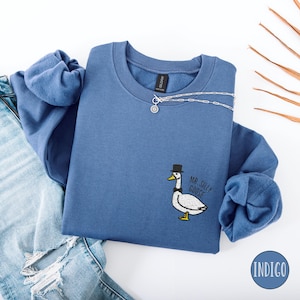 May include: A blue sweatshirt with a white goose wearing a top hat and the text "Mr. Silly Goose" embroidered on the front. The sweatshirt is laid out on a white surface with a pair of blue jeans.