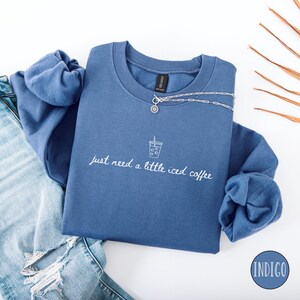 K&ouml;nnte beinhalten: Indigoblaues Sweatshirt mit dem Text "just need a little iced coffee" und einer Grafik eines Eiskaffees. Eine silberne Halskette mit Anh&auml;nger befindet sich auf dem Sweatshirt. Das Sweatshirt ist &uuml;ber einer Jeans gefaltet.