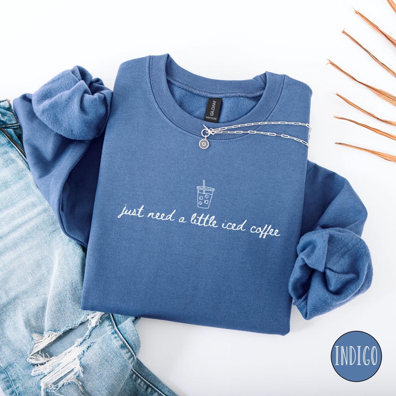 K&ouml;nnte beinhalten: Indigoblaues Sweatshirt mit dem Text "just need a little iced coffee" und einer Grafik eines Eiskaffees. Eine silberne Halskette mit Anh&auml;nger befindet sich auf dem Sweatshirt. Das Sweatshirt ist &uuml;ber einer Jeans gefaltet.