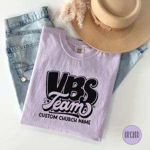 Op de afbeelding: Een lichtpaarse T-shirt met een zwarte afbeelding waarop "VBS Team" staat met sterren en de tekst "Custom Church Name".