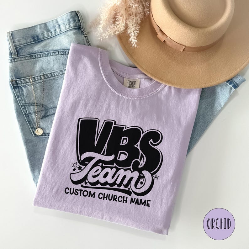 Op de afbeelding: Een lichtpaarse T-shirt met een zwarte afbeelding waarop "VBS Team" staat met sterren en de tekst "Custom Church Name".