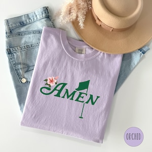 May include: A lavender t-shirt with a green golf flag and the word "AMEN" in a cursive font. A pink flower is above the word "AMEN".