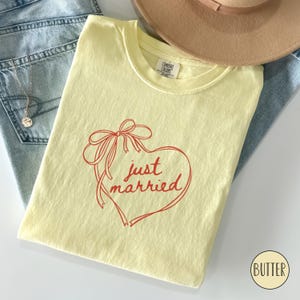 Op de afbeelding: Een botergeel T-shirt met een rood hartvormig ontwerp en de tekst "just married". Het shirt is opgevouwen en ligt op een blauwe spijkerbroek en een beige hoed. Een zilveren ketting is zichtbaar.
