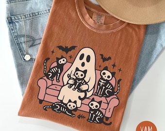May include: A burnt orange t-shirt featuring a Halloween-themed graphic. The design includes a ghost, skeleton cats, bats, and a pink couch. The shirt is paired with blue jeans and a tan hat.
