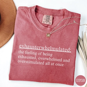 Op de afbeelding: Een maronkleurig T-shirt met witte tekst die "exhausterwhelmulated: the feeling of being exhausted, overwhelmed and overstimulated all at once" luidt.
