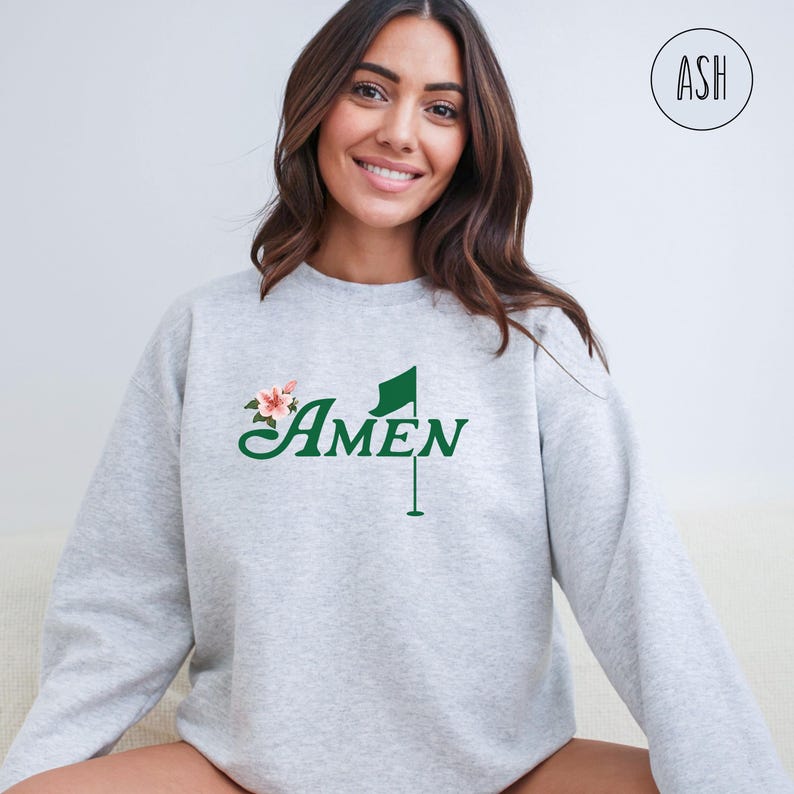 May include: A gray sweatshirt with a green golf flag and the word "AMEN" in a cursive font. There are pink flowers on the left side of the design.