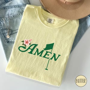 May include: A yellow t-shirt with a green golf flag and the word "AMEN" in green lettering. There is a pink flower graphic above the word "AMEN".