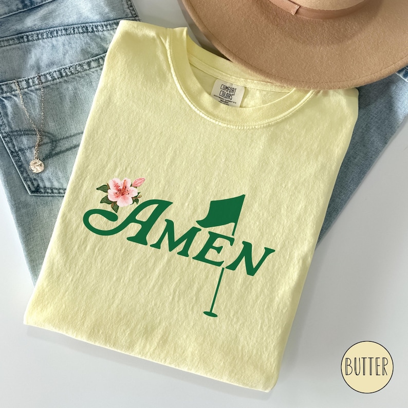 May include: A yellow t-shirt with a green golf flag and the word "AMEN" in green lettering. There is a pink flower graphic above the word "AMEN".