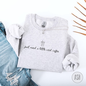 K&ouml;nnte beinhalten: Hellgraues Sweatshirt mit dem handgeschriebenen Text "just need a little iced coffee". Oberhalb des Textes befindet sich eine kleine Strichzeichnung einer Eiskaffeetasse. Eine silberne Halskette und eine blaue Jeans sind ebenfalls im Bild zu sehen.