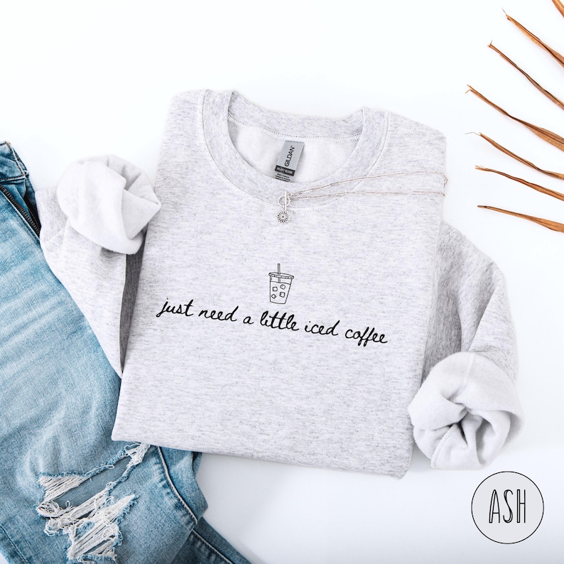 K&ouml;nnte beinhalten: Hellgraues Sweatshirt mit dem handgeschriebenen Text "just need a little iced coffee". Oberhalb des Textes befindet sich eine kleine Strichzeichnung einer Eiskaffeetasse. Eine silberne Halskette und eine blaue Jeans sind ebenfalls im Bild zu sehen.