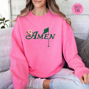 May include: A pink sweatshirt with a green golf flag and the word "AMEN" in a green font. The sweatshirt has a floral design with pink flowers.