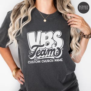 Op de afbeelding: Een donkergrijs T-shirt met een witte afbeelding waarop "VBS Team" staat, met daaronder een aangepaste kerknaam.