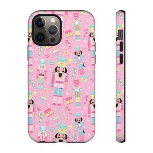 May include: A pink phone case featuring a repeating pattern of nutcrackers, candy canes, and cakes. The nutcrackers are in various colors, including pink, blue, and yellow. The case has a black border and a camera cutout.