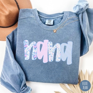 May include: A blue jean-colored sweatshirt with the word "nana" in a floral, gingham, and striped design. A gold necklace with a pendant is draped on the sweatshirt. A brown hat is visible in the background.