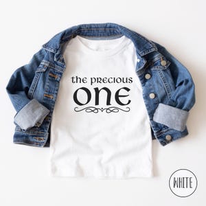 May include: White t-shirt with black text that reads "the precious one" with a decorative flourish underneath. The shirt is displayed on a denim jacket.