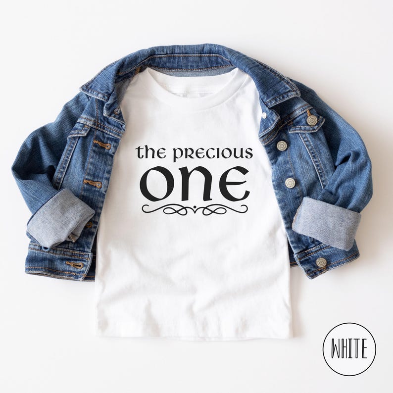 May include: White t-shirt with black text that reads "the precious one" with a decorative flourish underneath. The shirt is displayed on a denim jacket.