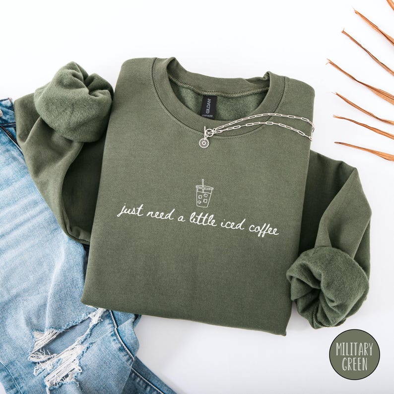 K&ouml;nnte beinhalten: Ein olivgr&uuml;nes Sweatshirt mit dem Text "just need a little iced coffee" und einer Eiskaffee-Grafik. Eine silberne Halskette liegt &uuml;ber der Vorderseite. Das Sweatshirt ist neben einer Jeans gefaltet.